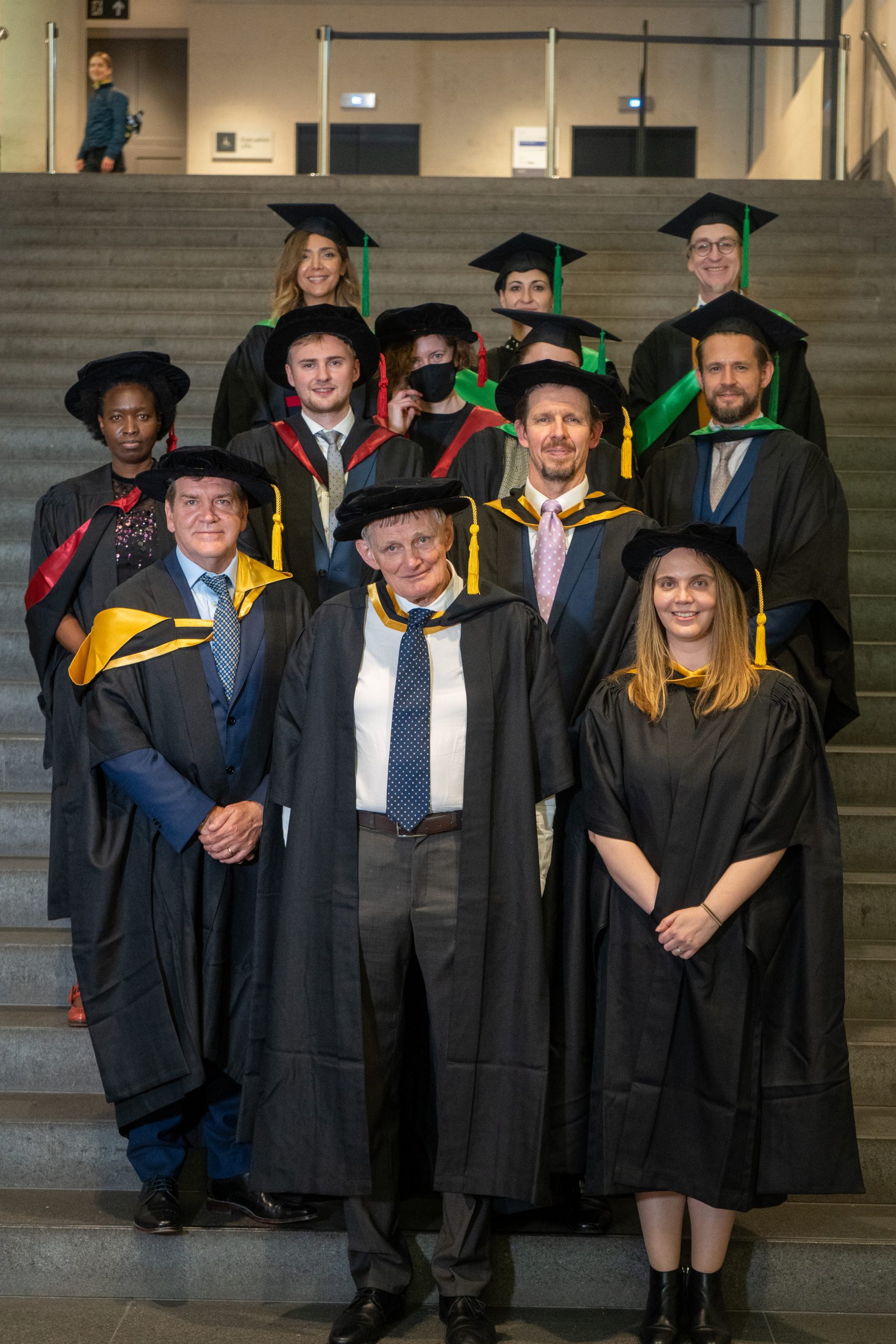 A group of academics dressed in graduation gowns pose for a picture. Each gown has a different colour to represent the different areas of DGHE: Business, Public Services, Health and Social Care and Art and Design.