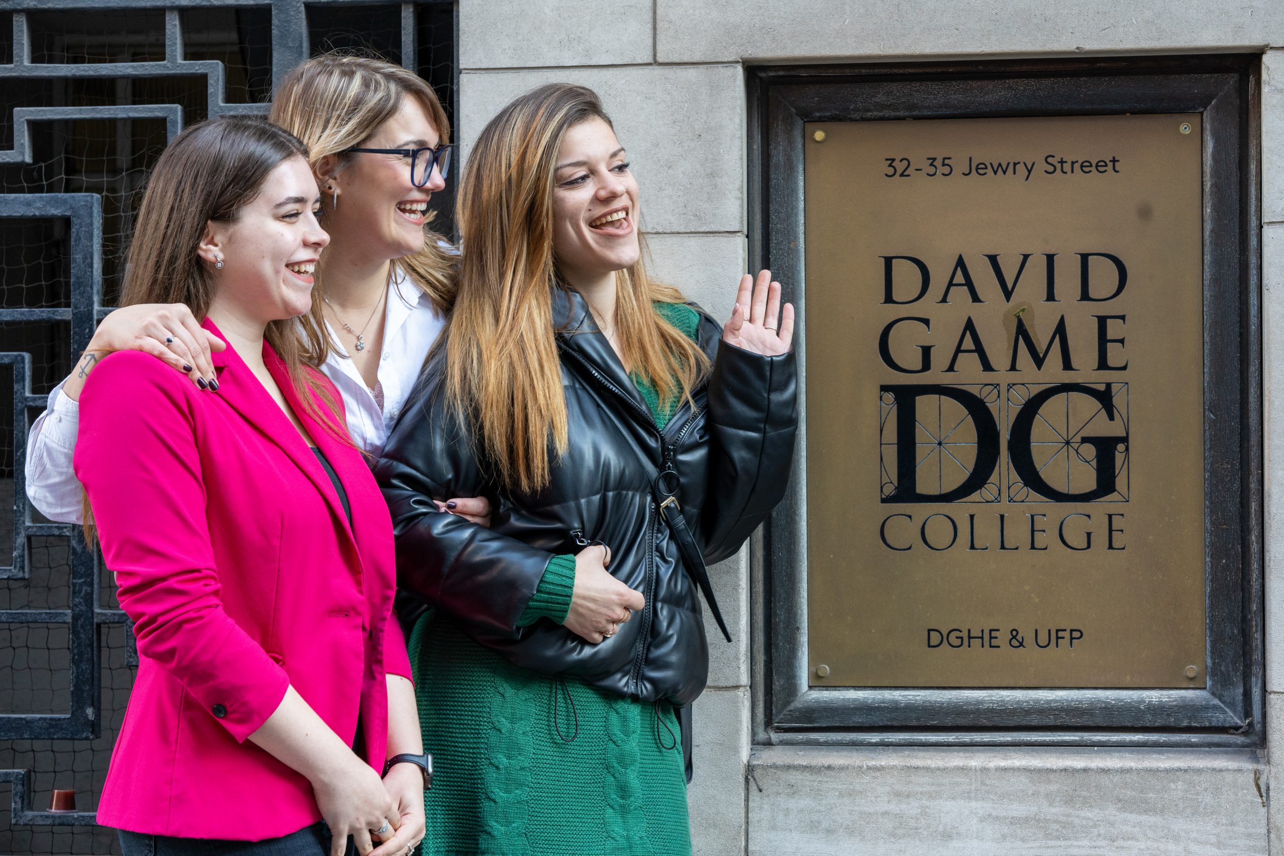 Three students stand next to the David Game College sign in front of the DGHE building. One of them waves at others out of the image. The women are very different from each other, and seem to be friends. They were casual clothing with bright colours.