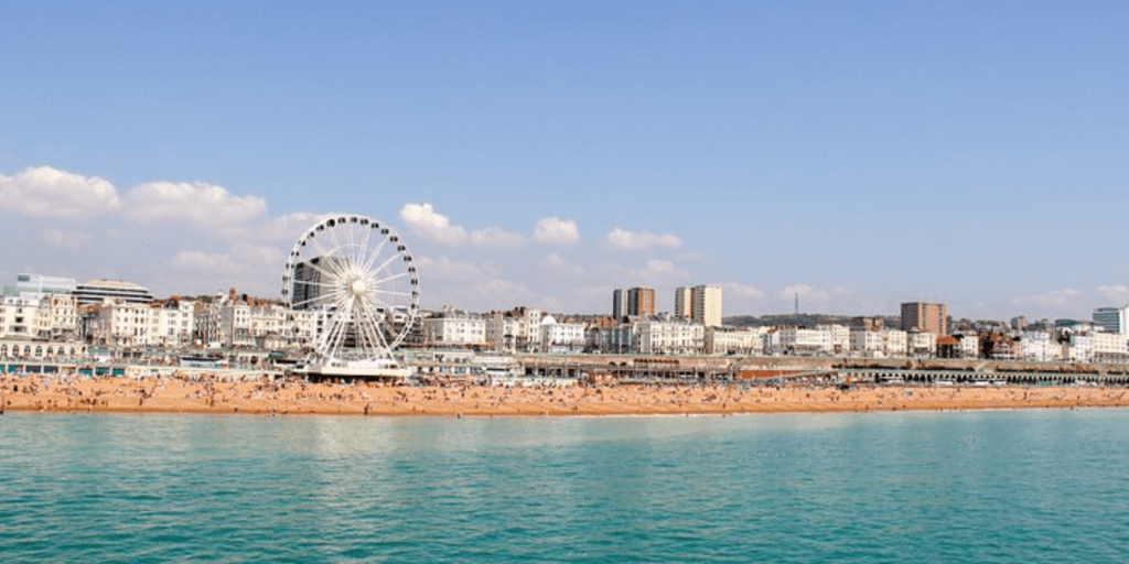 A picture of Brighton Beach seafront from the ocean. We can see the water and a big part of the beach full of people enjoying the summer. We can also see a clear sky and a Ferris wheel.