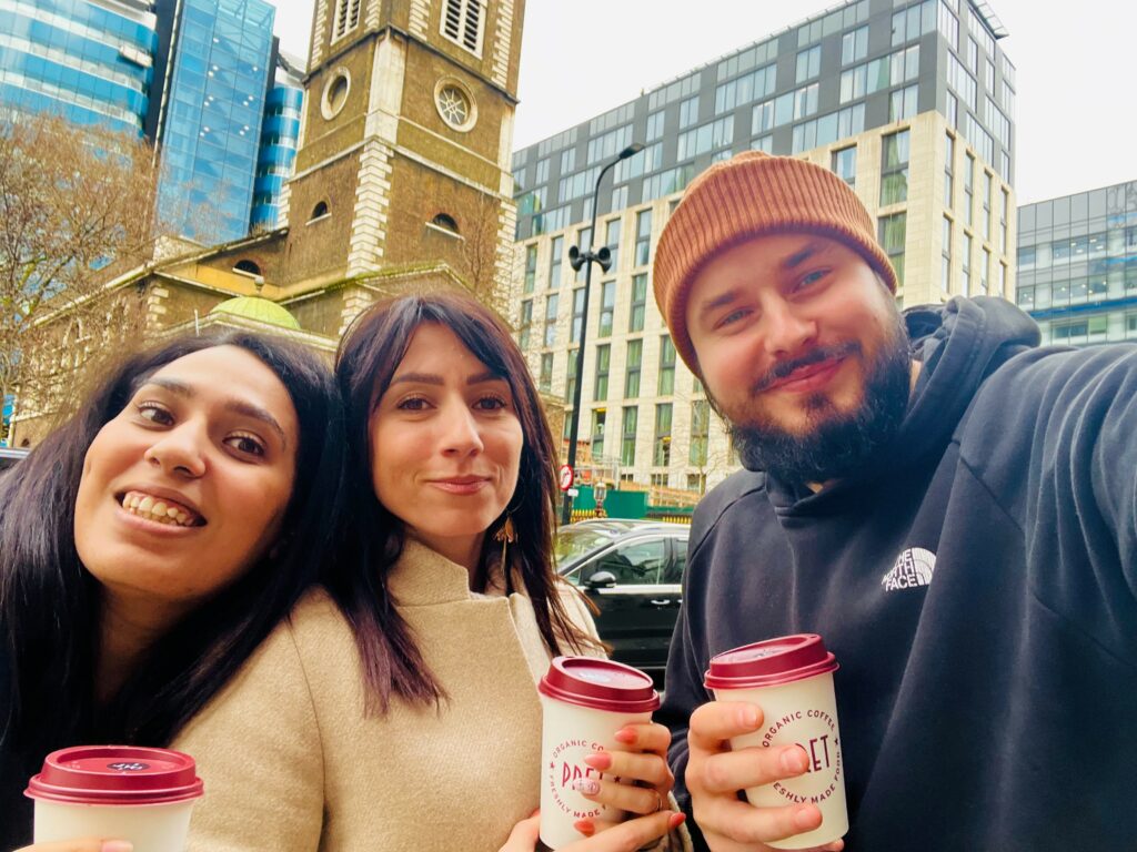 Three students gather in front of London landmarks with a coffee in hand.