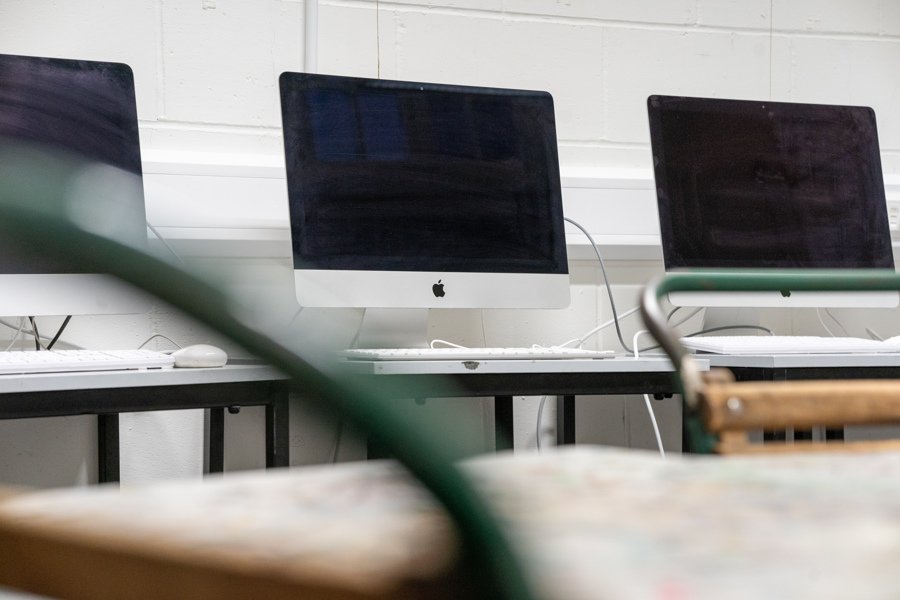 We see three Mac computers lined in front of a white, clean wall. You can also see a work station for art and design activities in the first area.