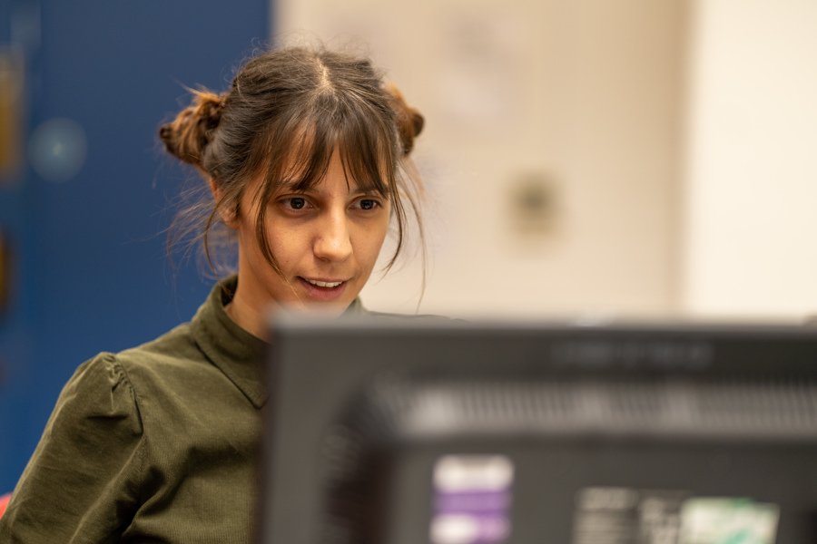 A young woman faces a computer in a computer lab. Her hair is in a bun and she seems concentrated on the activity.