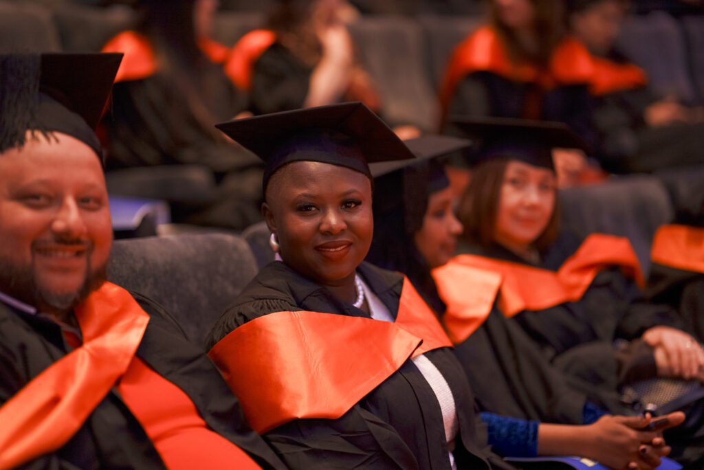 A group of graduates wearing caps and gowns with orange stoles, seated and smiling during a graduation ceremony.